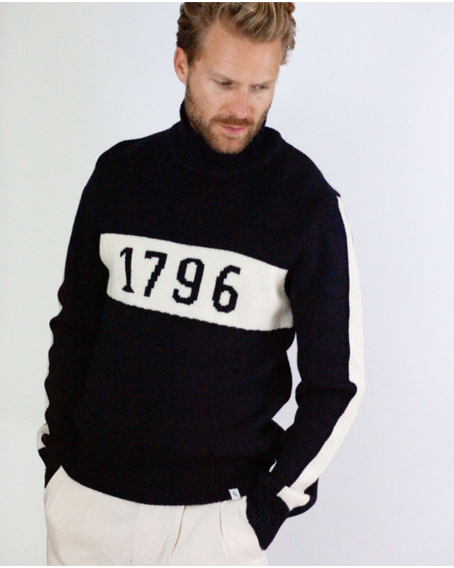 Flatlay image of 1796 navy merino wool knitted jumper. Navy jumper with white stripe across the chest detailing 1796 in navy 