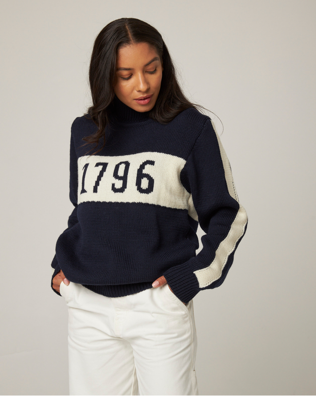 Flatlay image of 1796 navy merino wool knitted jumper. Navy jumper with white stripe across the chest detailing 1796 in navy 