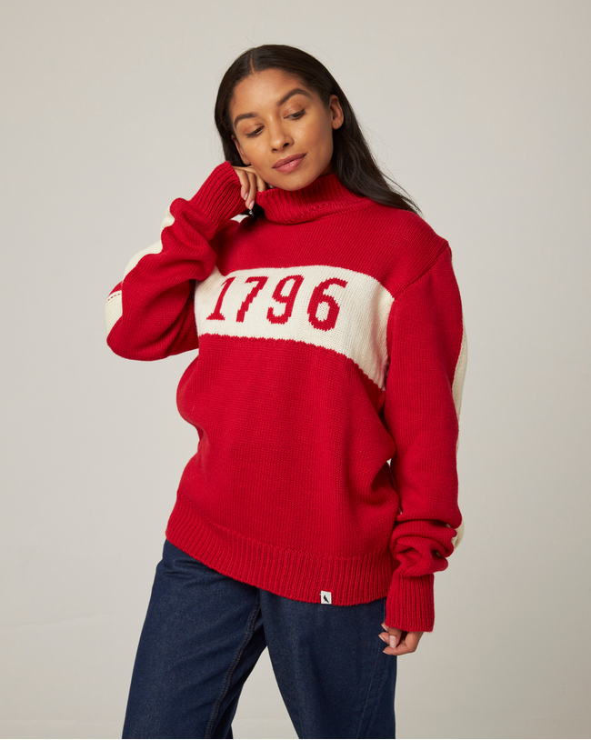 Flatlay image of 1796 red merino wool knitted jumper. red jumper with white stripe across the chest detailing 1796 in red. Woman wearing red jumper arms crossed across her chest. Woman wearing red jumper smiling crossing arms across her chest