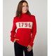 Flatlay image of 1796 red merino wool knitted jumper. red jumper with white stripe across the chest detailing 1796 in red. Woman wearing red jumper arms crossed across her chest. Woman wearing red jumper smiling crossing arms across her chest