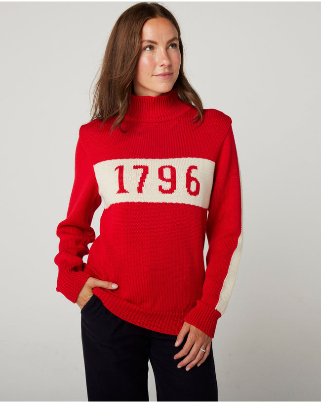 Flatlay image of 1796 red merino wool knitted jumper. red jumper with white stripe across the chest detailing 1796 in red. Woman wearing red jumper arms crossed across her chest. Woman wearing red jumper smiling crossing arms across her chest