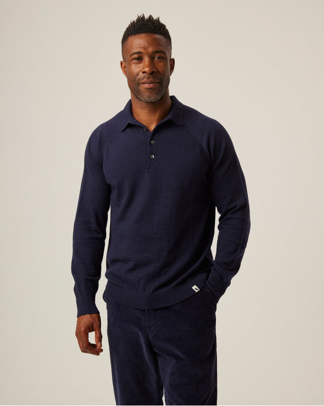 A flatlay image of a navy blue polo shirt jumper. The jumper has a collar and  4 buttons to the chest. The hem and cuff have a ribbed detail. The jumper is a light weight, fine knit jumper and only one colour all over. A male model wearing the jumper and looking of to the left. A male model wearing the jumper outside whilst walking across a wooden beam in a field. He is wearing the jumper with beige trousers and black shoes.