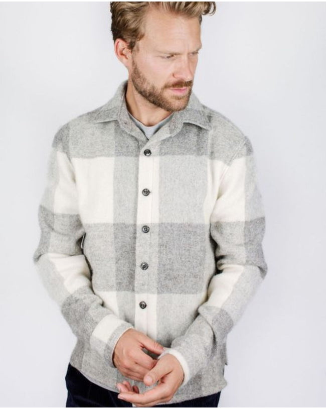 Flatlay image of a checked wool collared shirt consisting of a light grey, slightly darker grey and cream with seven small black buttons fastening down the center front fastening the shirt closed, the shirt has two discreet pockets on the either front bottom side of the shirt.