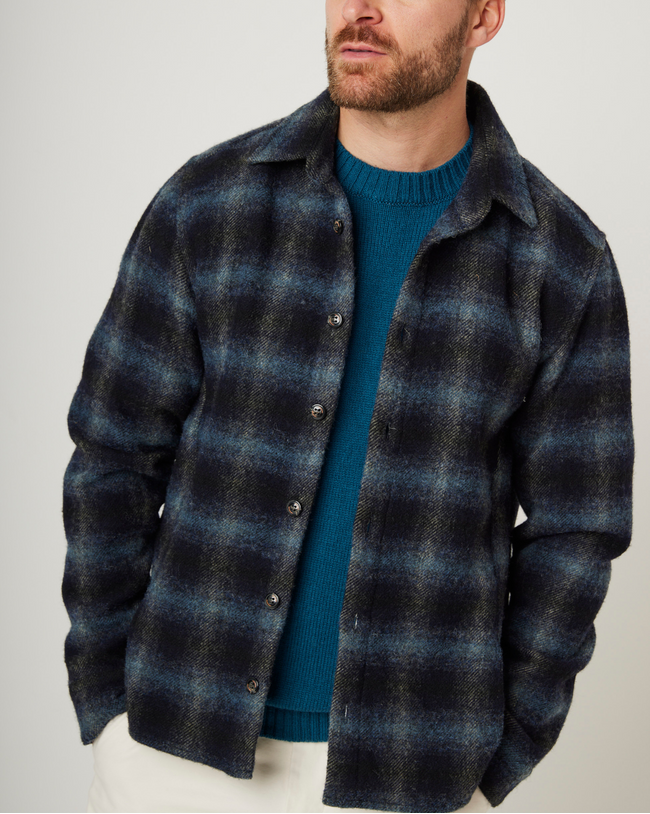 Flatlay image of a navy checked wool shirt with seven buttons down the center front fastening the collared shirt closed, the shirt has a slightly curved hem, the main base colour of the shirt is navy with the check pattern being a lighter shade of grey/blue.